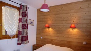 A bedroom with a wooden headboard, two bedside lamps, a bed with a white duvet, a window with patterned curtains, and a radiator.