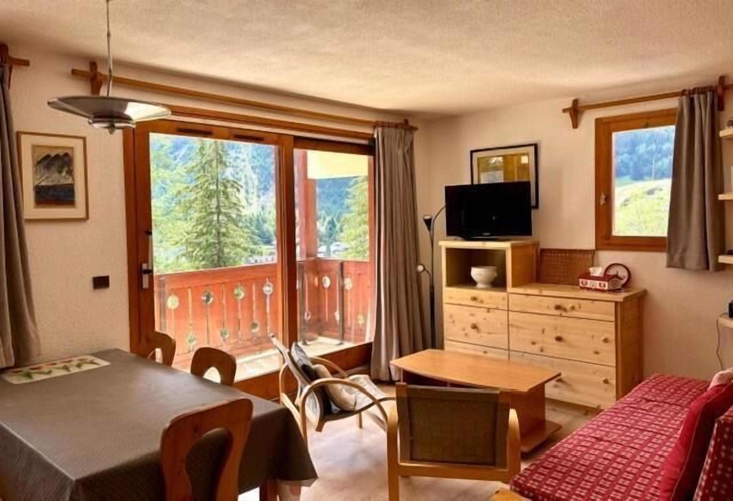 A room with a dining table, chairs, a television on a dresser, a bed with a red blanket, and a balcony with a view of trees.