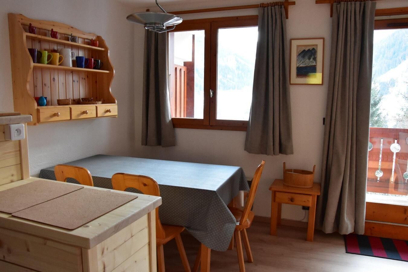 Una habitación acogedora con una mesa de madera, sillas, una estantería con tazas de colores, una ventana con cortinas y un cuadro en la pared.