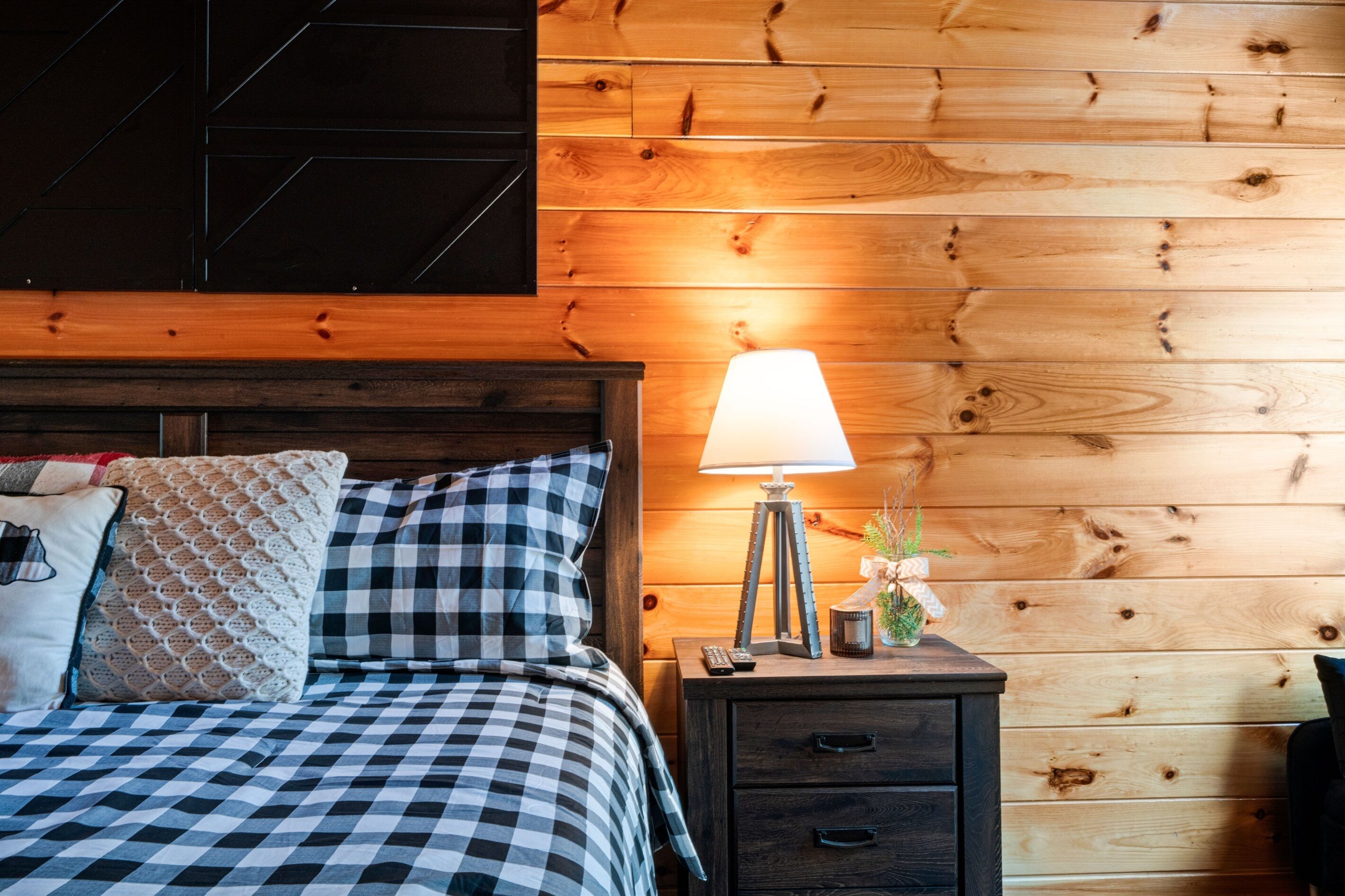 A bedroom with a checkered bedspread, a wooden headboard, a bedside lamp, and a nightstand with a remote control.