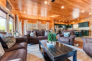 A cozy living room with a wooden ceiling, leather sofas, a coffee table, and a kitchen in the background.