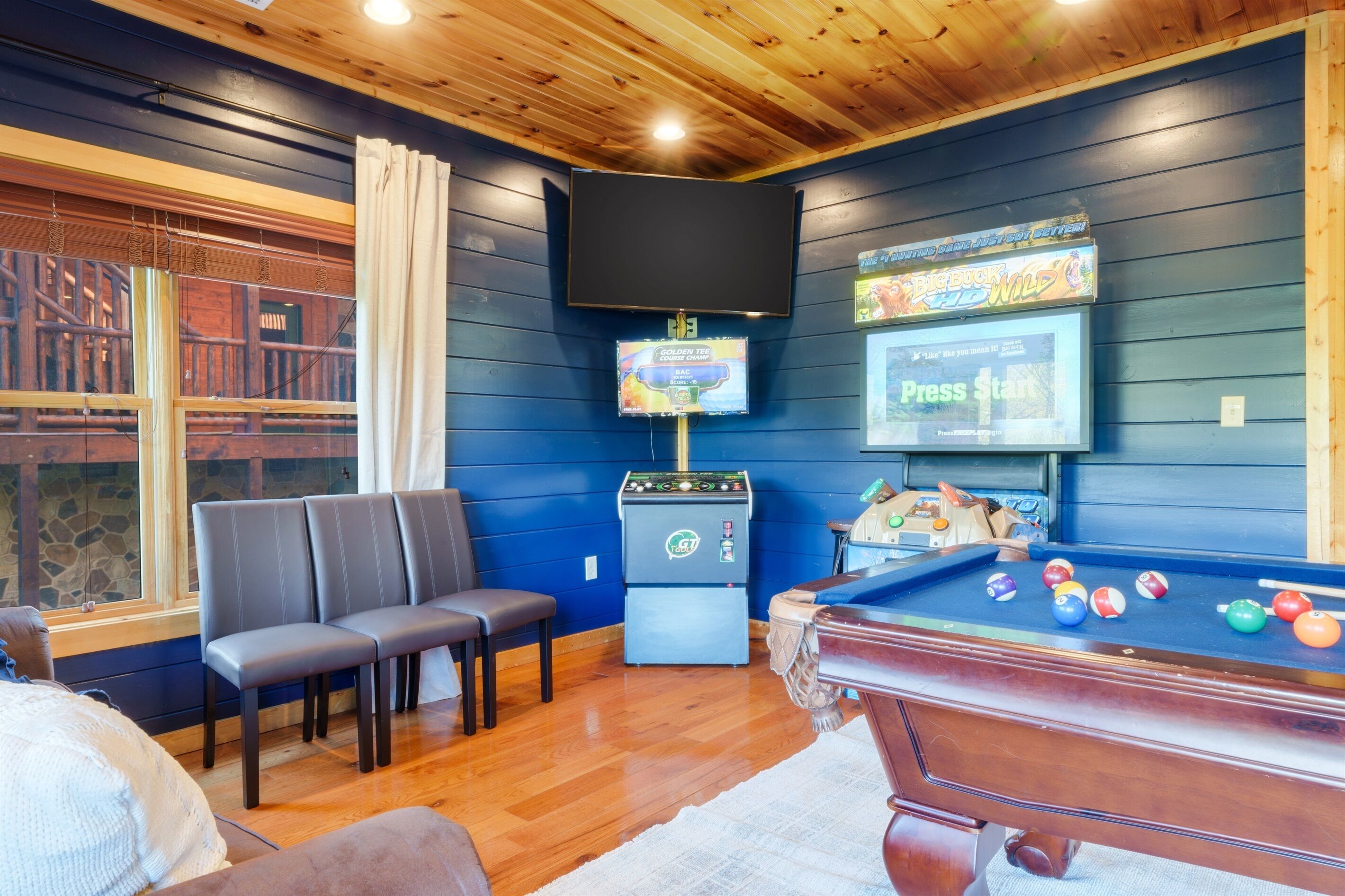 A pool table with balls, a television, and a jukebox in a room with wooden walls and a window.
