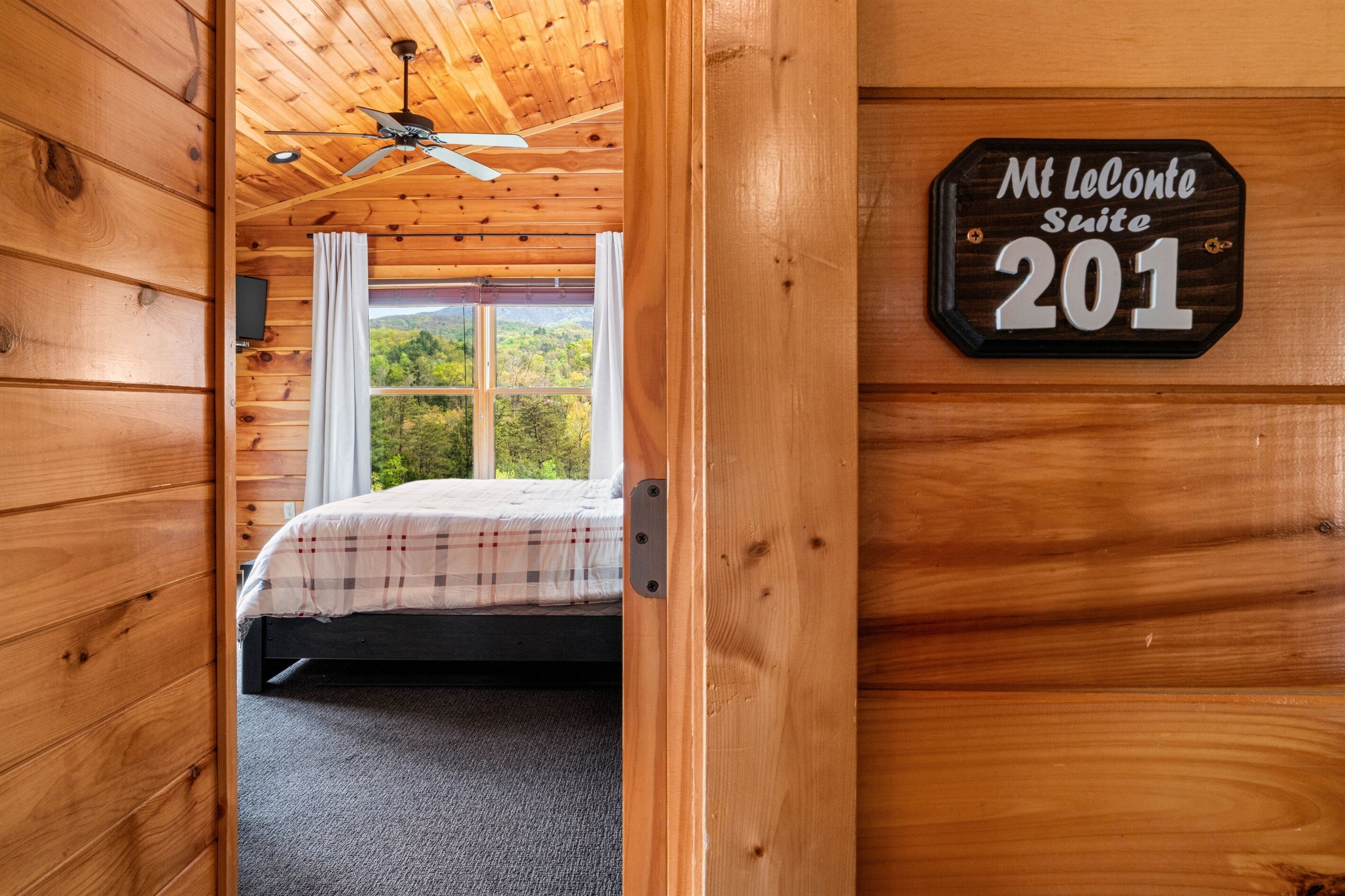 A wooden cabin interior with a bed, a ceiling fan, and a window with curtains. A sign on the wall reads 'Mt LeConte Suite 201'.