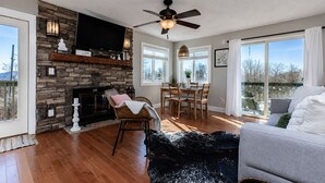 Sala de estar con chimenea de piedra, ventilador de techo, comedor con mesa y sillas, y ventanas grandes con vistas.