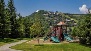Un parque infantil con toboganes y una estructura para escalar, rodeado de césped y árboles, con casas en una colina al fondo.