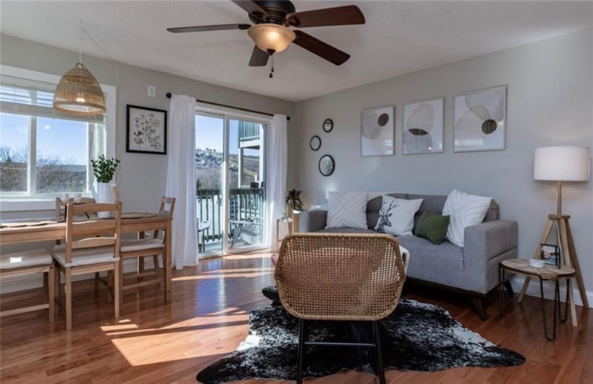 A modern living room with a ceiling fan, a grey sofa, a wicker chair, a dining table with chairs, and a large window with a view.