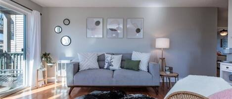 Un salon moderne avec un canapé gris, un tapis noir et un fauteuil en osier. Trois tableaux encadrés ornent le mur, une lampe se trouve sur une table d’appoint et une fenêtre est agrémentée de rideaux.