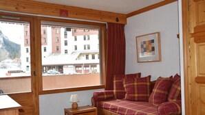 Ein gemütliches Zimmer mit einer Holzdirektiva, einem kleinen Tisch und einem Fenster mit Blick auf Gebäude und Berge.