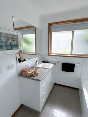 A modern bathroom with a white sink, mirror, and tiled floor. There is a window with frosted glass and a black towel hanging on a rack.