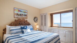 Un dormitorio con cama, ventana con vista al mar, un cuadro en la pared y una puerta con contraventanas.