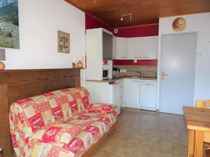 A cozy living area with a sofa and a wooden table, a kitchen with white cabinets, and a door with a clock.
