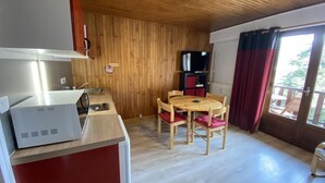 Ett kompakt kök med en mikrovågsugn, ett matbord i trä med fyra stolar, en tv och ett fönster med gardiner.