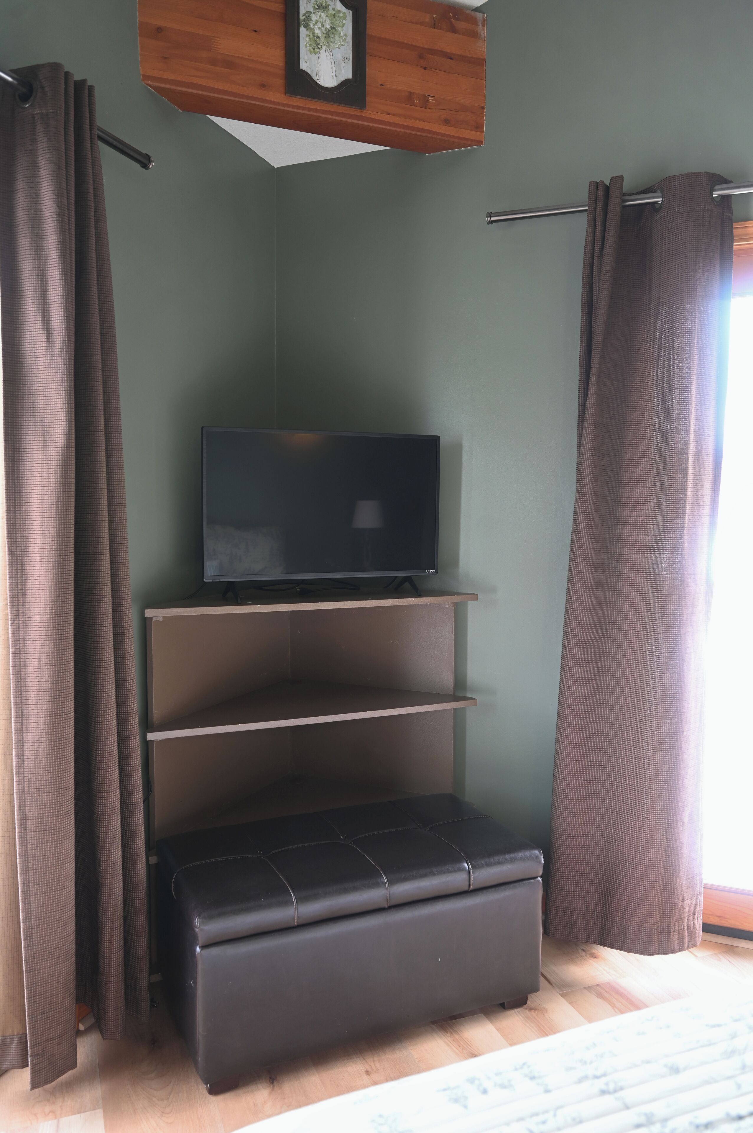 Une pièce avec un téléviseur posé sur une étagère, un pouf noir et des rideaux aux fenêtres.
