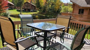 Una terraza con una mesa y sillas de vidrio, rodeada por una valla de madera, con vistas a un estanque y árboles de colores otoñales.