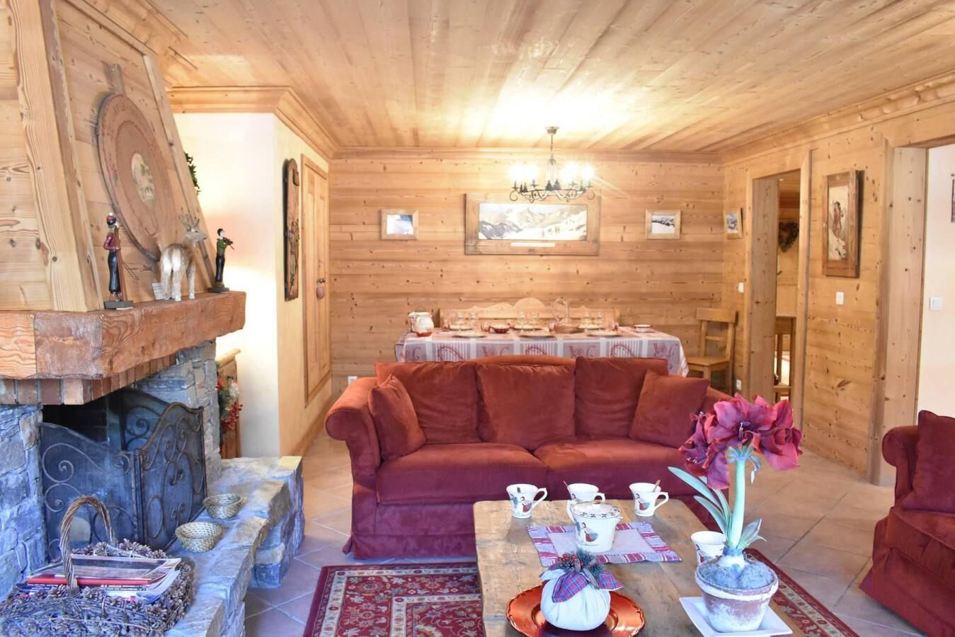 Interior acollidor d'una cabana de fusta amb una llar de foc, un sofà vermell, una taula de dinar preparada per a quatre i una làmpada penjant.