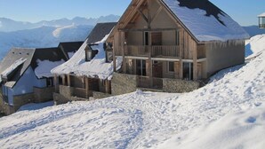 A chalet-style mountain lodge with a wooden exterior and stone foundation, surrounded by snow-covered terrain and distant mountains.
