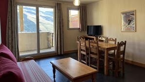 A living room with a sofa, a wooden table, and a TV. There are curtains on the windows, and a poster on the wall.