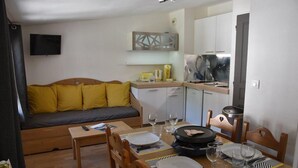 A compact living space with a sofa, dining table, chairs, a kitchen area, and a TV mounted on the wall.
