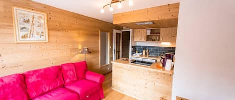 A cozy room with a wooden floor, a bright red sofa, a wooden dining table, and a kitchen area with wooden cabinets and a tiled backsplash.