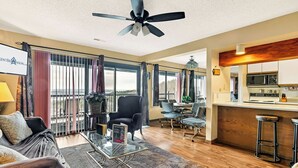 A spacious living room with a large window, a ceiling fan, a dining area, and a kitchen with bar stools.