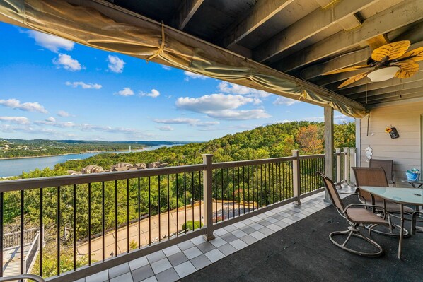 Ein Balkon mit Blick auf einen See und einen Wald, ein Tisch mit Stühlen und ein Deckenventilator.