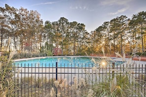 A swimming pool surrounded by a black metal fence, with tall trees in the background and a clear sky.
