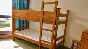 Lliri amb lliteres de fusta i roba de llit blanca en una habitació amb finestra, un tamboret i un petit armari.