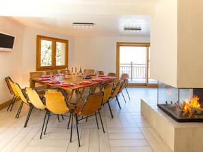 Un coin repas avec une table ronde dressée pour six personnes, entourée de chaises, d’une cheminée et d’une grande fenêtre donnant sur un beau paysage.