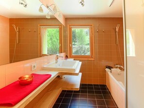Une salle de bain avec deux lavabos, une baignoire et une douche. Des fenêtres laissent entrer la lumière naturelle et offrent une vue sur la verdure environnante.