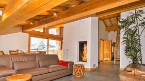 A cozy living room with a fireplace, wooden beams, a sofa, and a dining area visible through a large window. - Chalet, wifi, sauna, terrace, balcony, fireplace or stove, parking, tv, 250m², Les Arcs (Bourg-Saint-Maurice)