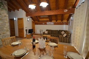 Uma área de refeições com uma mesa de madeira posta para quatro pessoas, uma televisão ao fundo e um sofá na sala de estar adjacente.