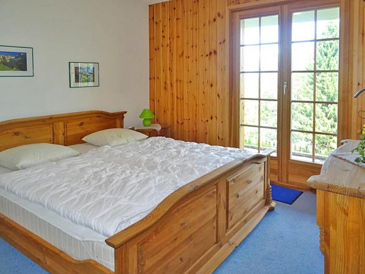 A wooden bed with a white mattress and headboard, a nightstand with a lamp, and a window with a view of greenery.