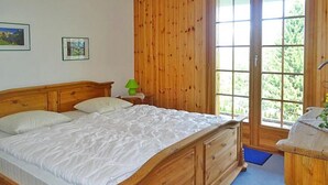 Una cama de madera con colchón blanco y cabecera, una mesita de noche con lámpara, y una ventana que da a zonas verdes.