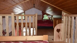 Une chambre mansardée en bois, avec un lit, une fenêtre ornée d’un rideau rouge et une porte en bois avec une découpe en forme de cœur.