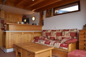 A cozy cabin interior with a wooden kitchenette, a sofa with patterned cushions, and a window with curtains. - 150m from resort center, 1st floor, balcony, tv, ski locker, 60m², Méribel (Méribel - Les Allues)