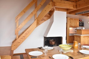 Comedor con mesa de madera dispuesta para cuatro, chimenea y cocina al fondo.