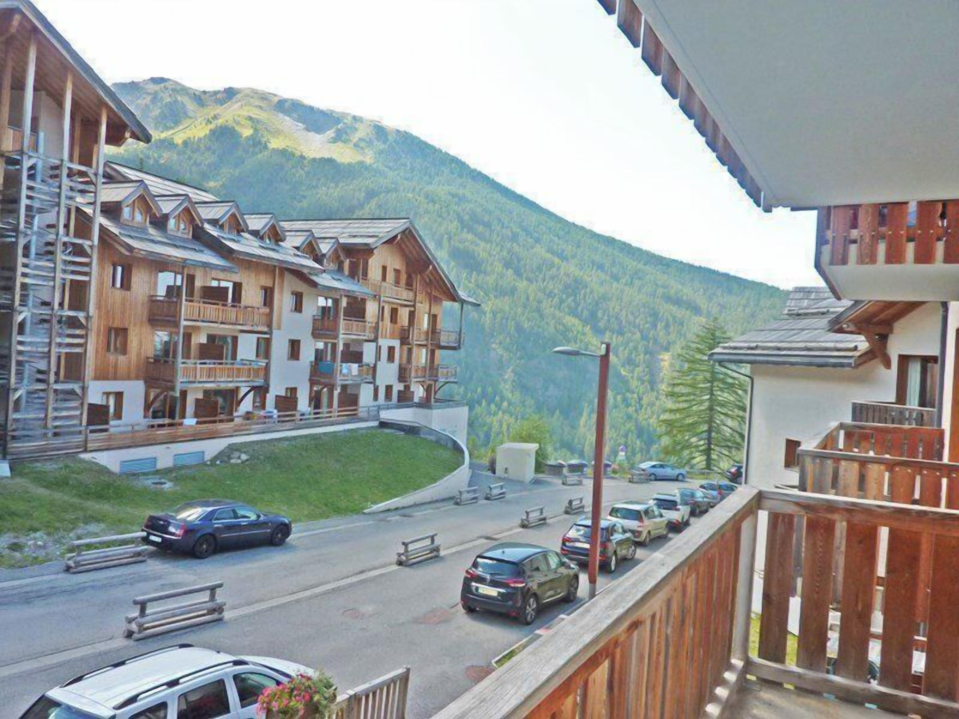 A multi-story wooden building with balconies, a parking area with cars, and a mountain in the background.