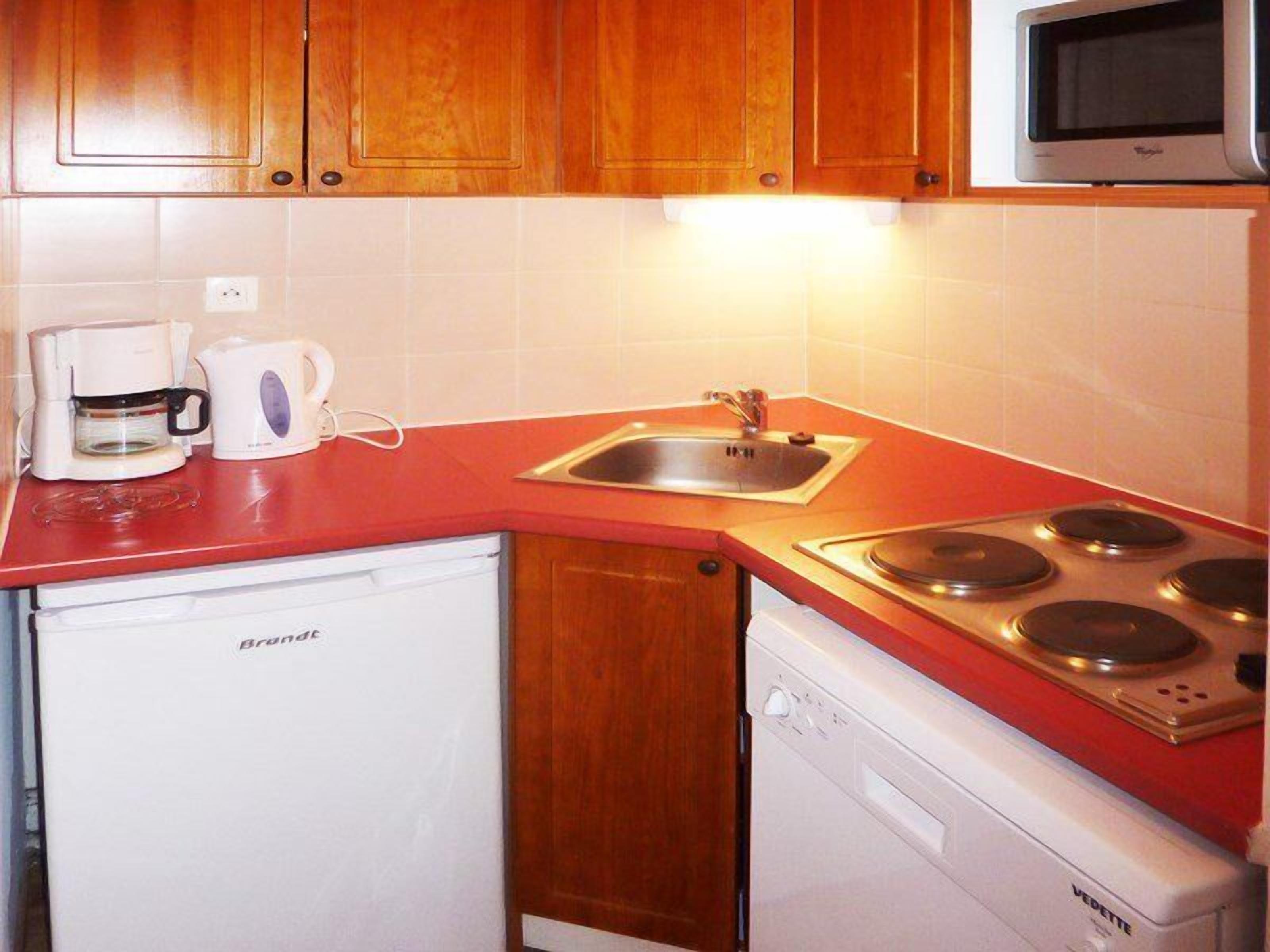 A compact kitchen with wooden cabinets, a red countertop, a built-in microwave, a coffee maker, a sink, and a dishwasher.