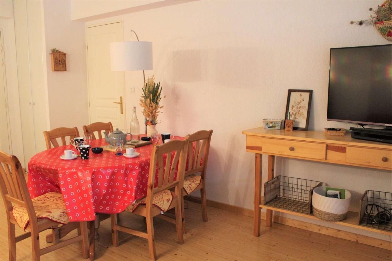 Et spiseområde med rødt borddække, træstole, en lampe, et fjernsyn og et lille skab.
