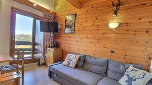 A cozy cabin room with a sofa, a television, a wooden cabinet, and a window with a view of the mountains. - 10m from ski slopes, 3rd floor, balcony, ski locker, 41m², Méribel-Mottaret (Méribel - Les Allues)