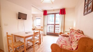 A cozy room with a wooden table and chairs, a bed with patterned bedding, a TV mounted on the wall, and large windows with curtains. - 130m from ski slopes, 1st floor, balcony, parking, tv, ski locker, 27m², La Norma (La Norma)
