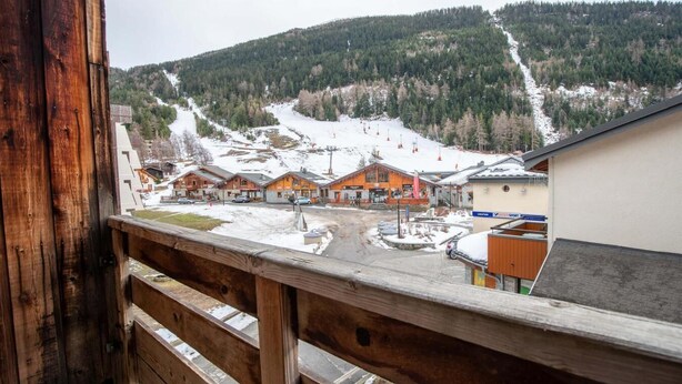 A wooden balcony overlooking a snow-covered village with rustic buildings and a ski slope in the background. - 130m from ski slopes, 1st floor, balcony, parking, tv, ski locker, 27m², La Norma (La Norma)