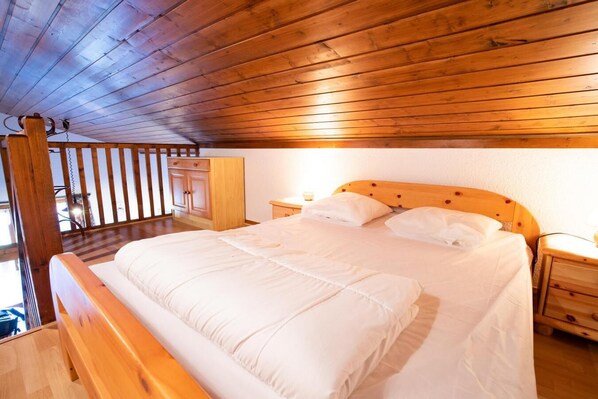 Un dormitorio con techo de madera, una cama con ropa de cama blanca, mesitas de noche de madera y un armario de madera.