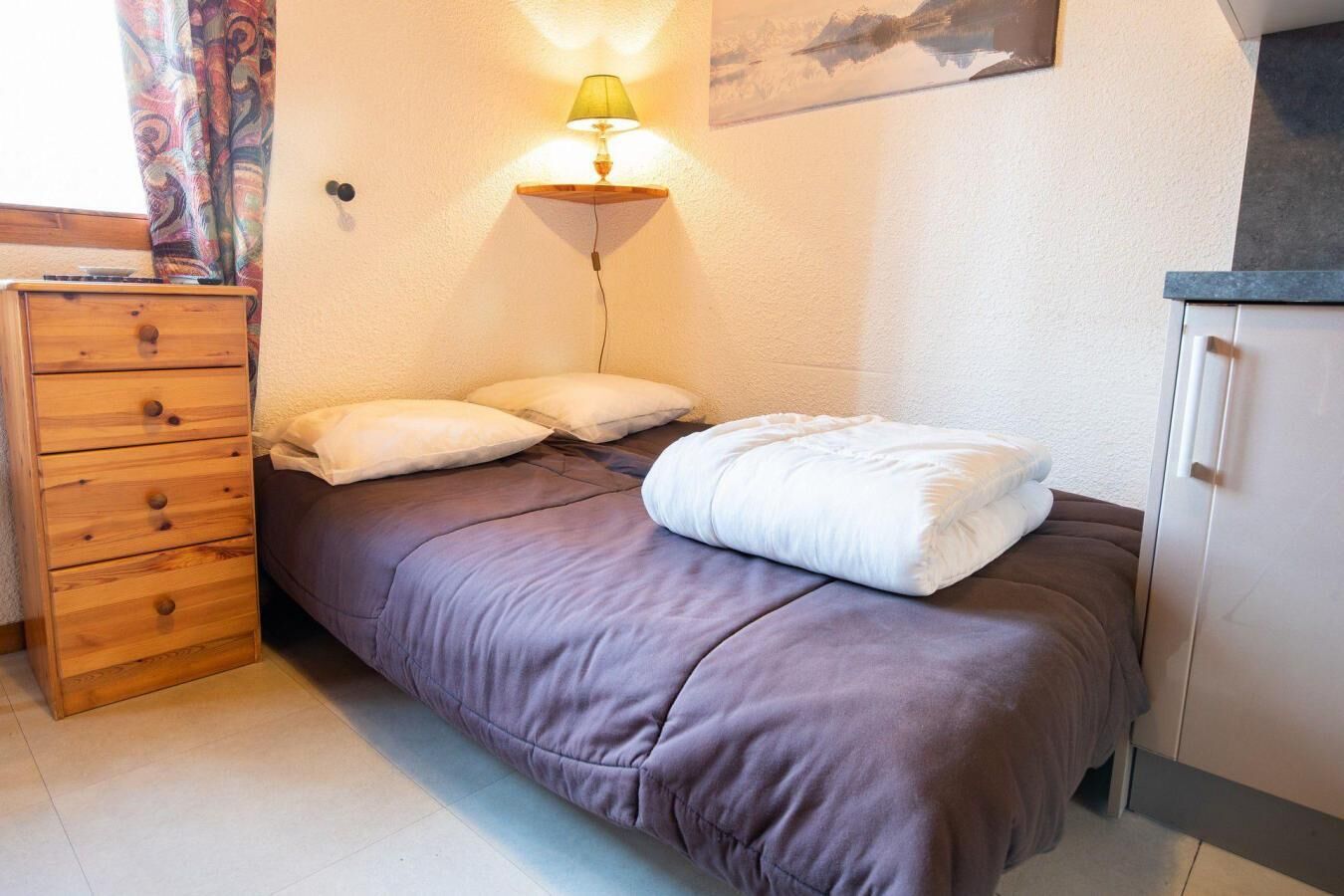 A single bed with a headboard, a bedside table with a lamp, a wooden dresser, and a cabinet.