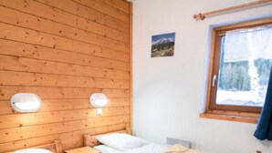 Et værelse med to køjesenge af træ, hver med en hvid madras og et beige tæppe. To vægmonterede lamper hænger over sengene, og et vindue viser udsigt til træer og bjerge.
