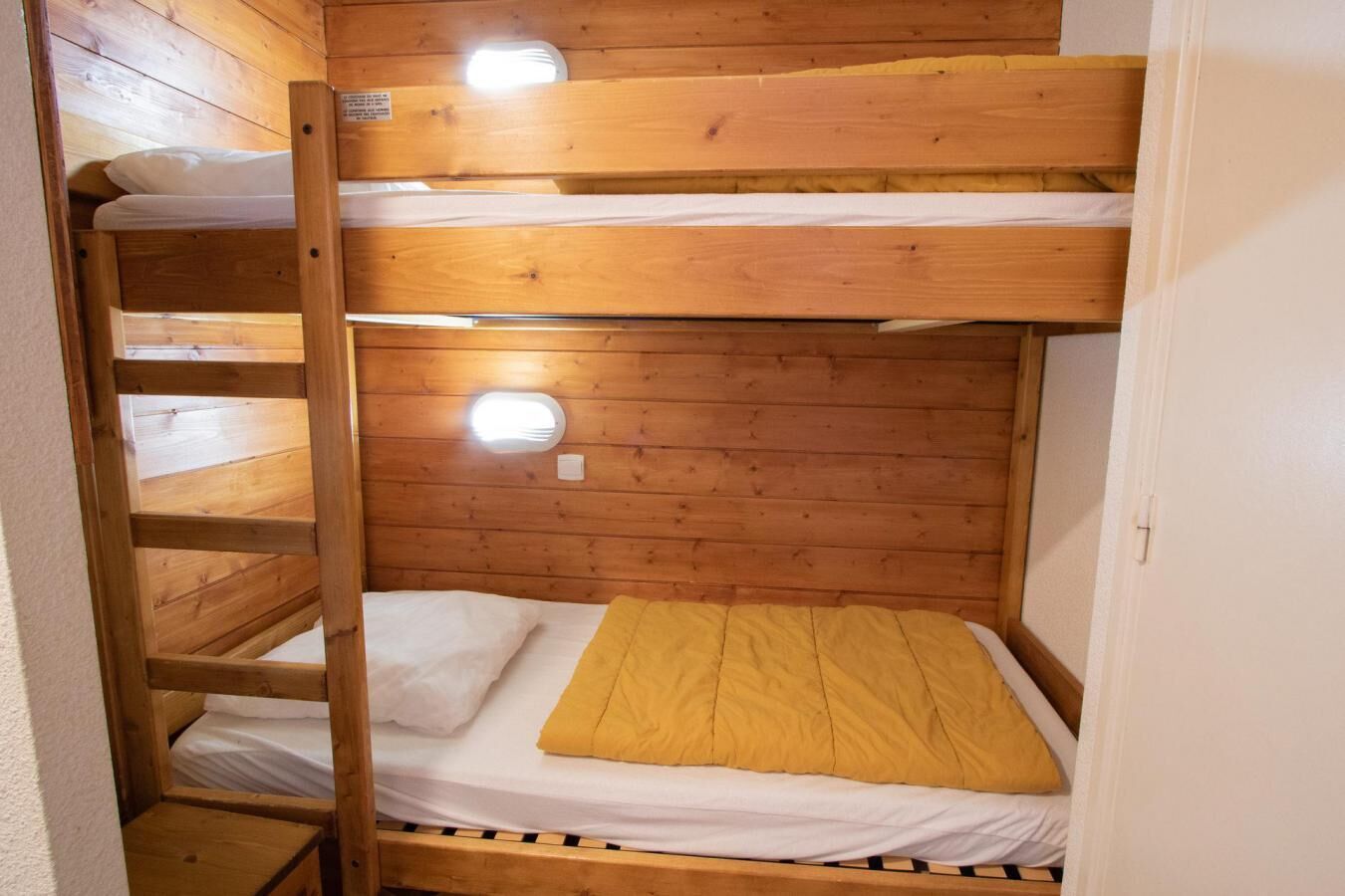 A bunk bed with a ladder, wooden walls, and a small window with a light fixture.