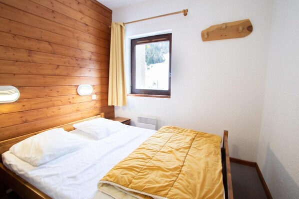 Ein Zimmer mit Holzwand, einem Bett mit weißer und gelber Bettwäsche, einem Fenster mit Blick auf Bäume und einer Wandleuchte.