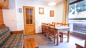 Une pièce chaleureuse avec un canapé à carreaux, une table à manger en bois et des chaises. On peut voir une porte en bois et une fenêtre donnant sur une zone boisée.