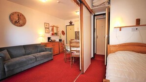 Una habitación acogedora con un sofá gris, una cama de madera, una alfombra roja, un armario de madera, una pequeña cocina y un reloj de pared.