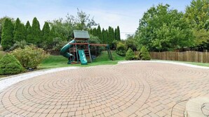 Een speeltuin met een glijbaan en schommels, omgeven door een rond pad van baksteen en begroeiing.